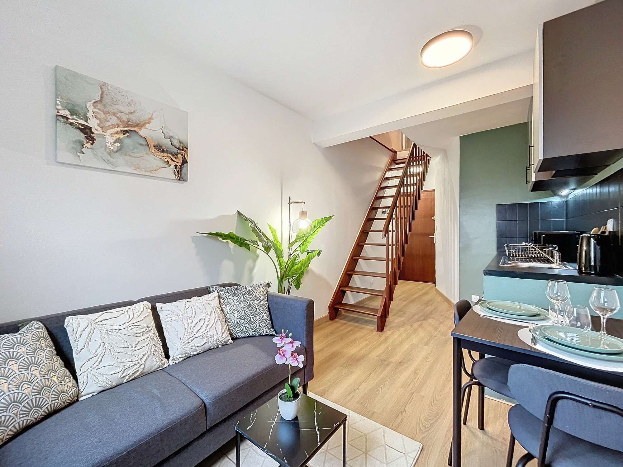 Ein modernes Wohnzimmer mit einem grauen Sofa, einem Esstisch für zwei Personen und einer Holztreppe, die in ein Obergeschoss führt.