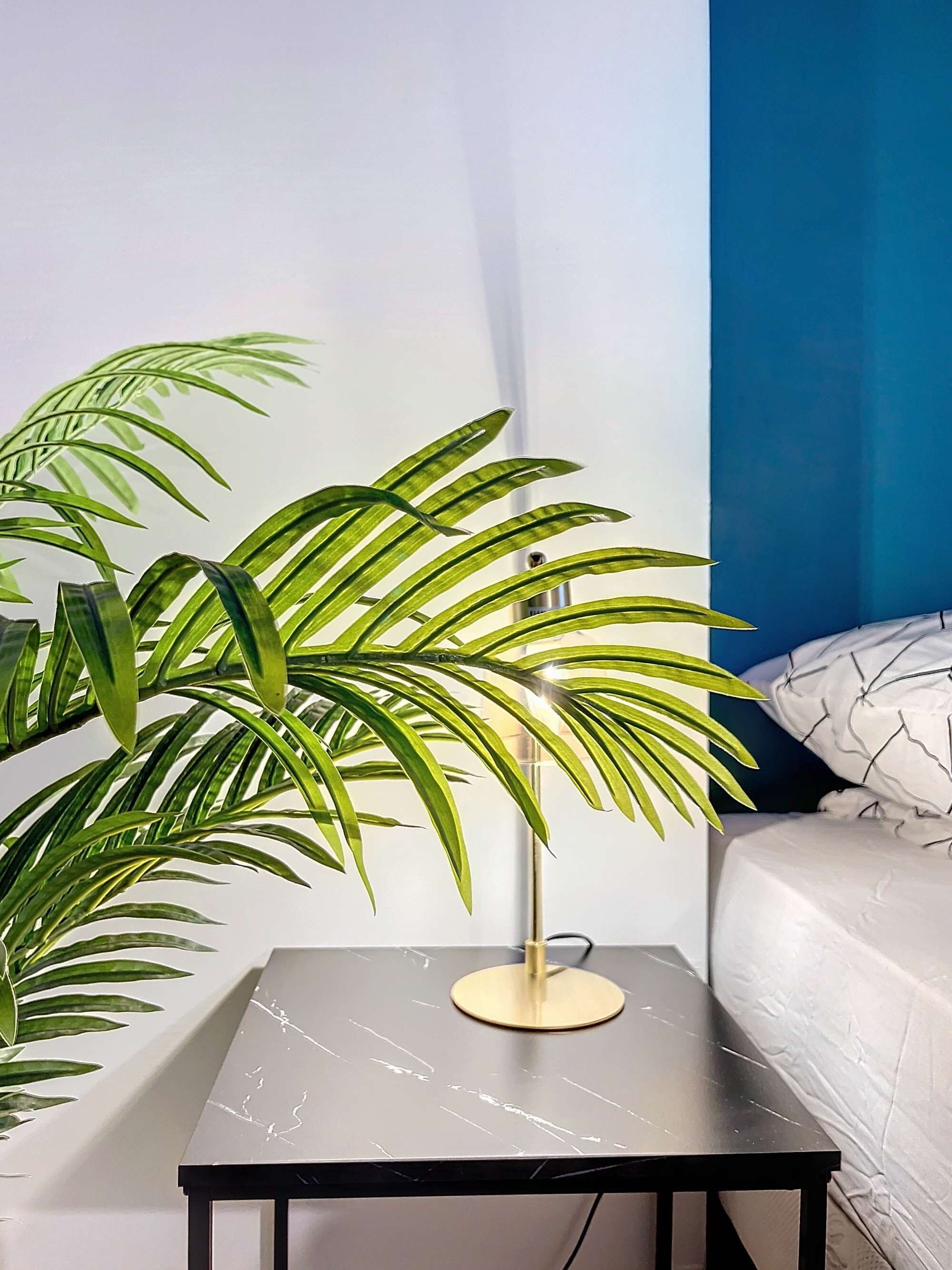 A small table with a modern lamp and a potted plant in a room with a bed and a blue wall.