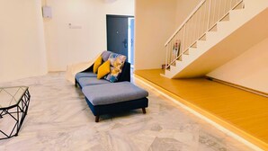 A modern living room with a sofa, ottoman, and a glass coffee table. A staircase with a wooden finish is visible to the right. - Indahpura Kulai (Kulai)