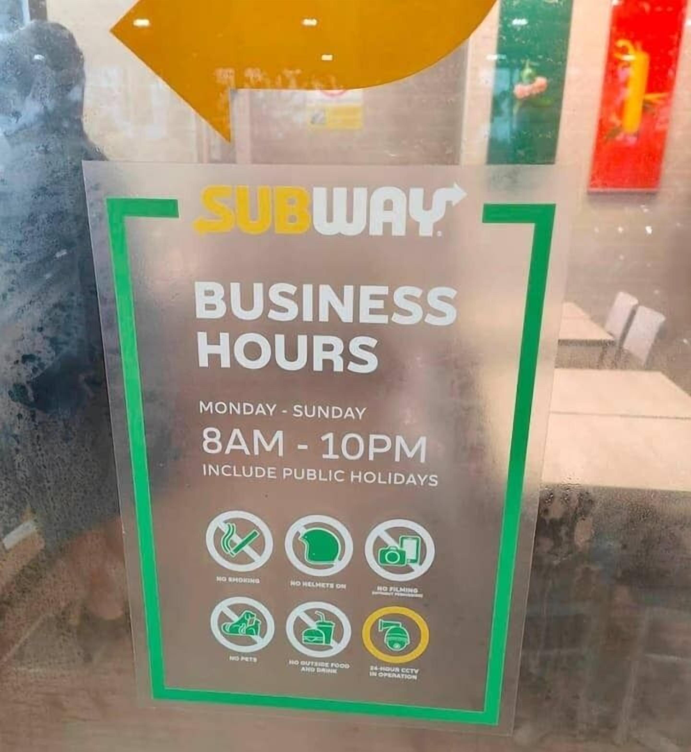 Schild der Subway mit Angabe der Öffnungszeiten, umrahmt von Grün, mit Symbolen für Rauchverbot, Verbot von Helmen, Verbot von Filmaufnahmen, Verbot von Haustieren, Verbot von Speisen und Getränken von außen sowie Hinweis auf eine 24-stündige Videoüberwachung.
