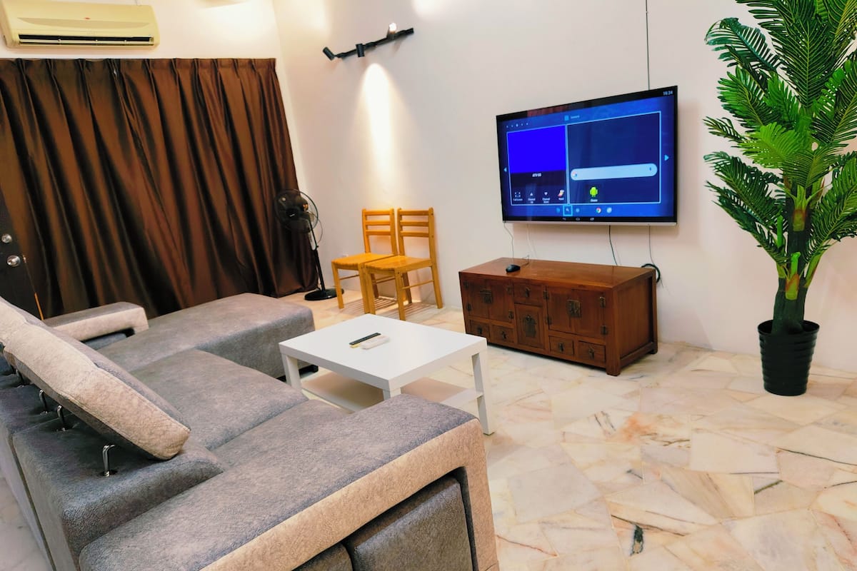 Een woonkamer met een bank, een televisie, een houten kast, een plant in pot en een ventilator.