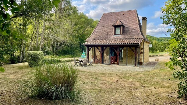 Una casa rústica con tejado de baldosas, un porche cubierto con mobiliario exterior y un jardín con diversas plantas y árboles.