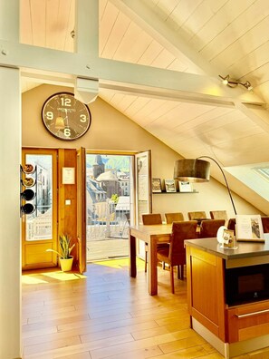 A hotel room with a wooden ceiling, a dining area with a table and chairs, a clock on the wall, and a view of the outside.