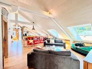 A spacious loft with a high ceiling, wooden beams, a cozy gray sofa, a dining area with a red sign, and a view through a skylight.