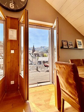 A room with a wooden floor, a dining table with chairs, a clock on the wall, and a view of a town with buildings and mountains.