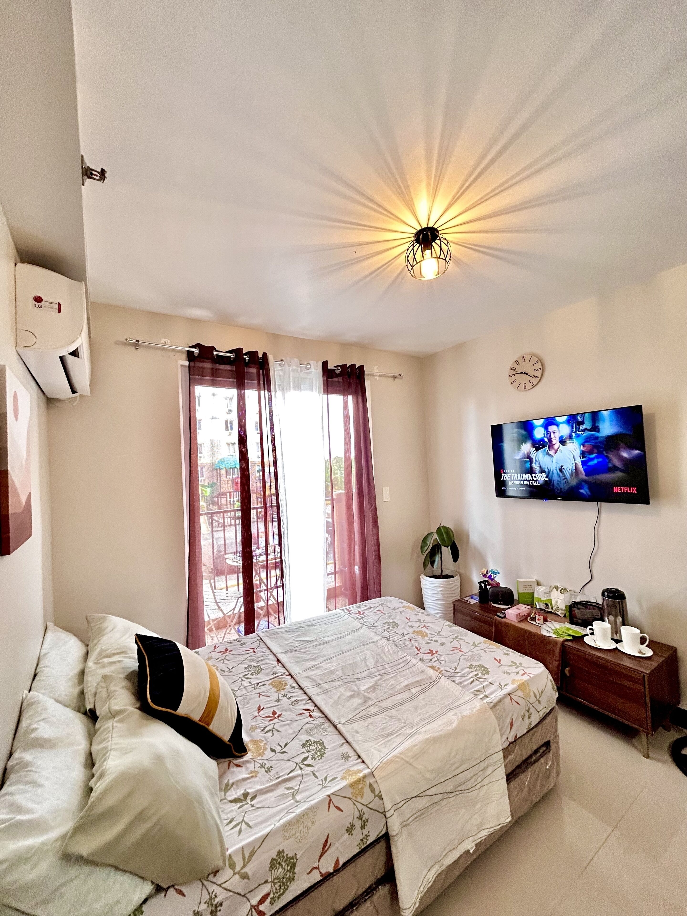 A hotel room with a bed, a TV mounted on the wall, a ceiling light fixture, and a window with curtains.