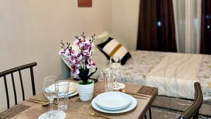 Un comedor ordenado con una mesa lista para dos, una orquídea en maceta y una cama con colcha blanca y marrón al fondo.