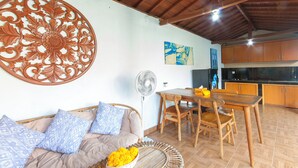 A living room with a sofa, a dining table, and chairs. There is a fan, a decorative wall piece, and a kitchen area with cabinets. - Tranquil Maha Villa Ubud (Ubud)