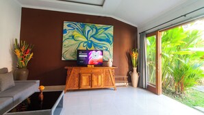 A living room with a flat-screen TV, a sofa, a coffee table, and a large window with a view of greenery. - Tranquil Maha Villa Ubud (Ubud)