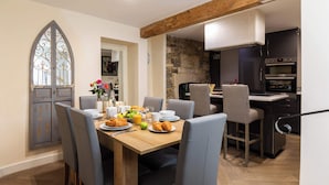A dining area with a wooden table, grey upholstered chairs, and a breakfast setting including croissants, fruit, and juice. A stone wall and modern kitchen with dark cabinets and stainless steel appliances are visible in the background.