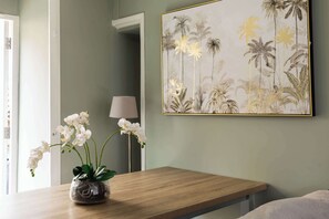 A room with a wooden table, a vase with white flowers, and a framed tropical plant artwork on the wall.