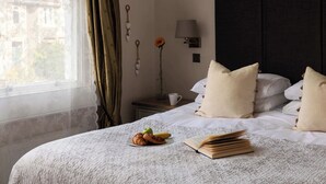 A neatly made bed with pillows, a bedside table with a lamp, a vase with a flower, and a plate with food.
