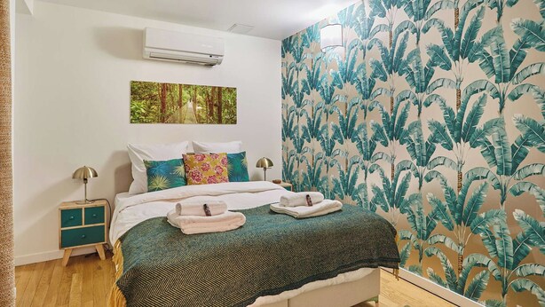 A bedroom with a bed, a nightstand, an air conditioner, a wall with a tropical leaf pattern, and a framed picture. - Charming Duplex Le Marais/beaubourg (Paris)