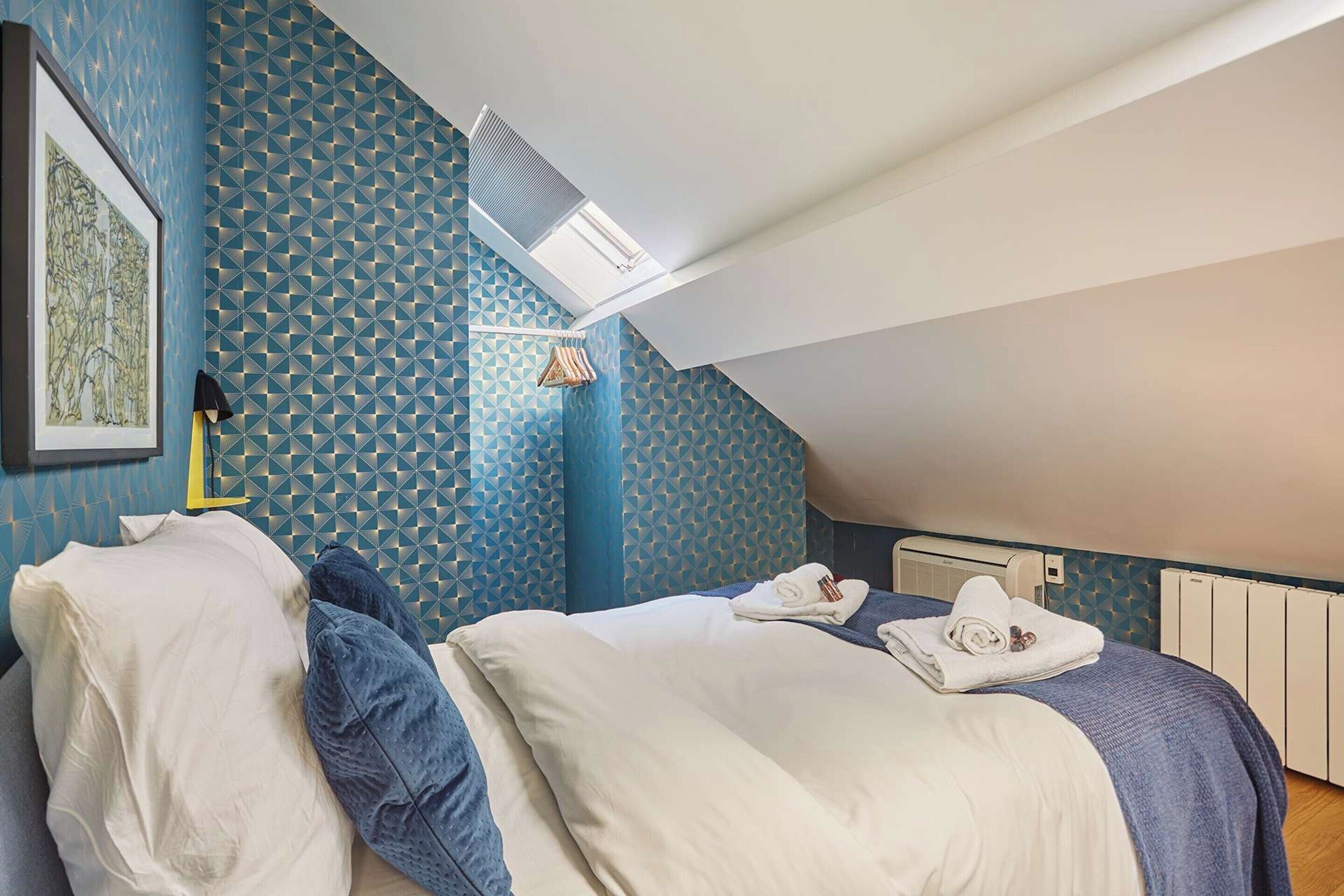 A bedroom with a sloped ceiling, a skylight, a bed with white and blue bedding, a framed picture on the wall, and a radiator.