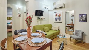 A dining area with a wooden table set for four, a green sofa, a grey armchair, and a TV mounted on the wall. - Charming Duplex Le Marais/beaubourg (Paris)