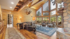 Un salón espacioso con pisos de madera, chimenea de piedra y grandes ventanales con vistas al exterior.