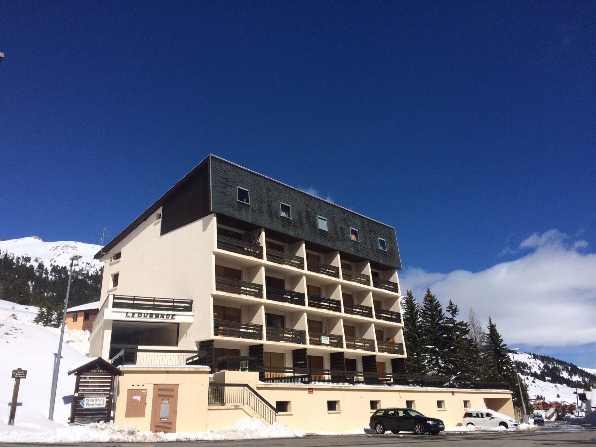 A multi-story hotel with a modern design, featuring balconies on each floor, a parking area, and a snowy mountain backdrop.