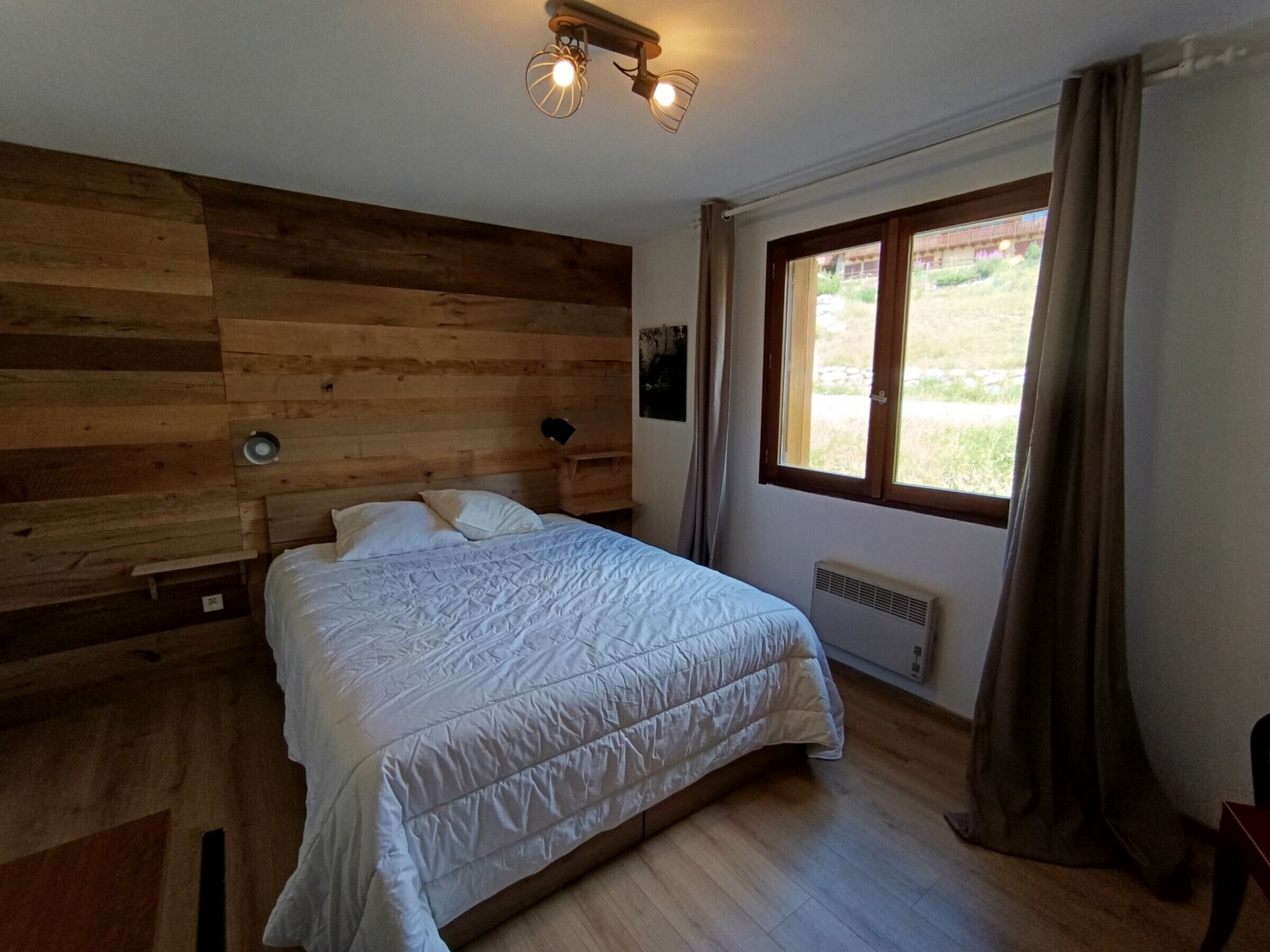A bedroom with a wooden headboard, a large bed with white bedding, a window with curtains, and a ceiling light fixture.