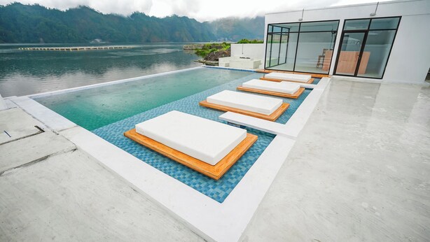 An infinity pool with white lounge chairs, a modern building with glass doors, and a mountainous backdrop. - Lake Batur Villa (Kintamani)
