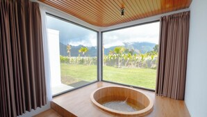 A modern bathroom with a wooden bathtub, large windows, and a view of a garden and mountains. - Lake Batur Villa (Kintamani)