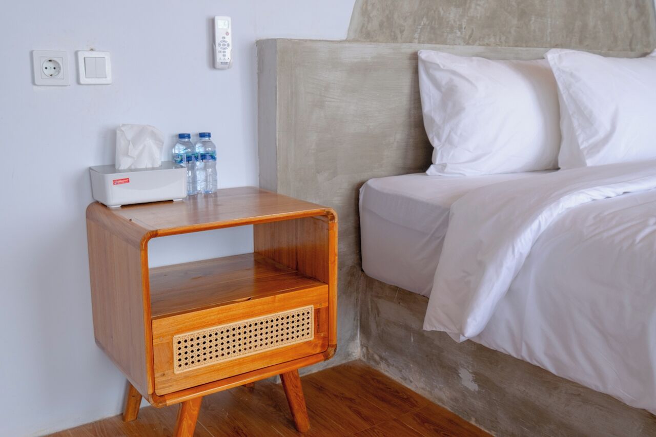 Een net opgemaakt bed met witte lakens, een nachtkastje met zakdoekjes en waterflesjes, en een stopcontact aan de muur.