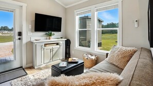 Ein Wohnzimmer mit einer Couch, einem Fernseher, einer Küchenzeile und großen Fenstern mit Blick auf ein Haus und Grünflächen.