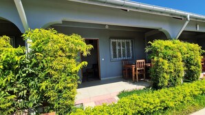 A covered outdoor area with a table and chairs, surrounded by greenery and a window with decorative bars. - Jardin fleuri de Lety (Nosy Be)