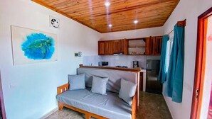 A cozy room with a wooden ceiling, a gray sofa, a kitchen area with wooden cabinets, and a framed painting of a turtle. - Jardin fleuri de Lety (Nosy Be)