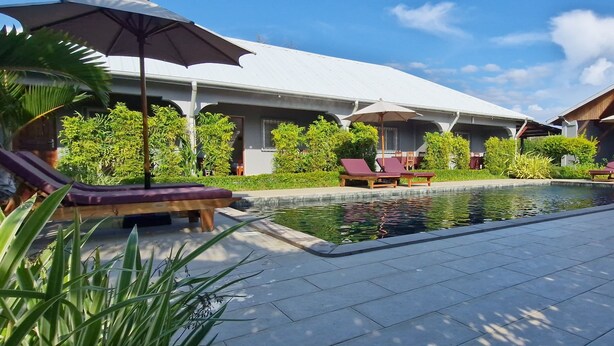 A poolside area with lounge chairs and umbrellas, surrounded by greenery and a building with a white roof. - Jardin fleuri de Lety (Nosy Be)