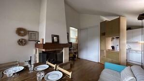 A dining area with a wooden table set for four, a fireplace, and a kitchen in the background. - Cozy apartment for 6 in Montgenèvre, pet-friendly with parking (Montgenèvre)
