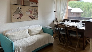 A cozy room with a sofa, dining table, and chairs. A large painting adorns the wall, and a window offers a view of trees and a building. - Cozy apartment for 6 in Montgenèvre, pet-friendly with parking (Montgenèvre)
