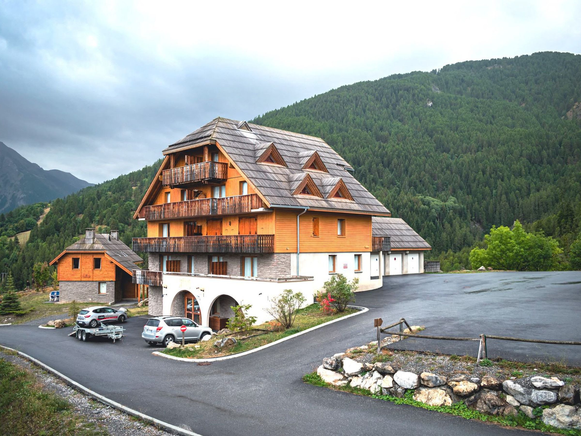 A multi-story wooden building with balconies, a stone foundation, and a parking area with cars.