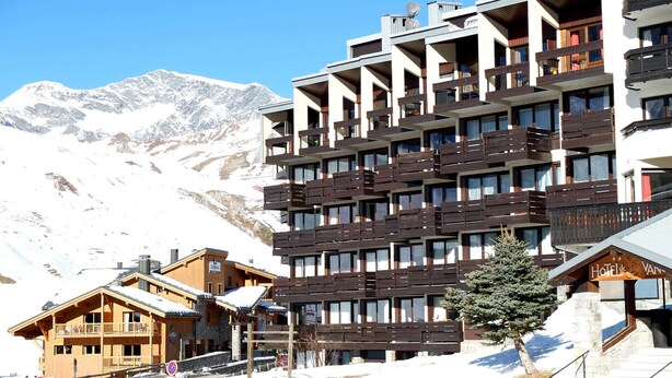A multi-story hotel with balconies, a ski slope in the background, and a mountainous landscape.