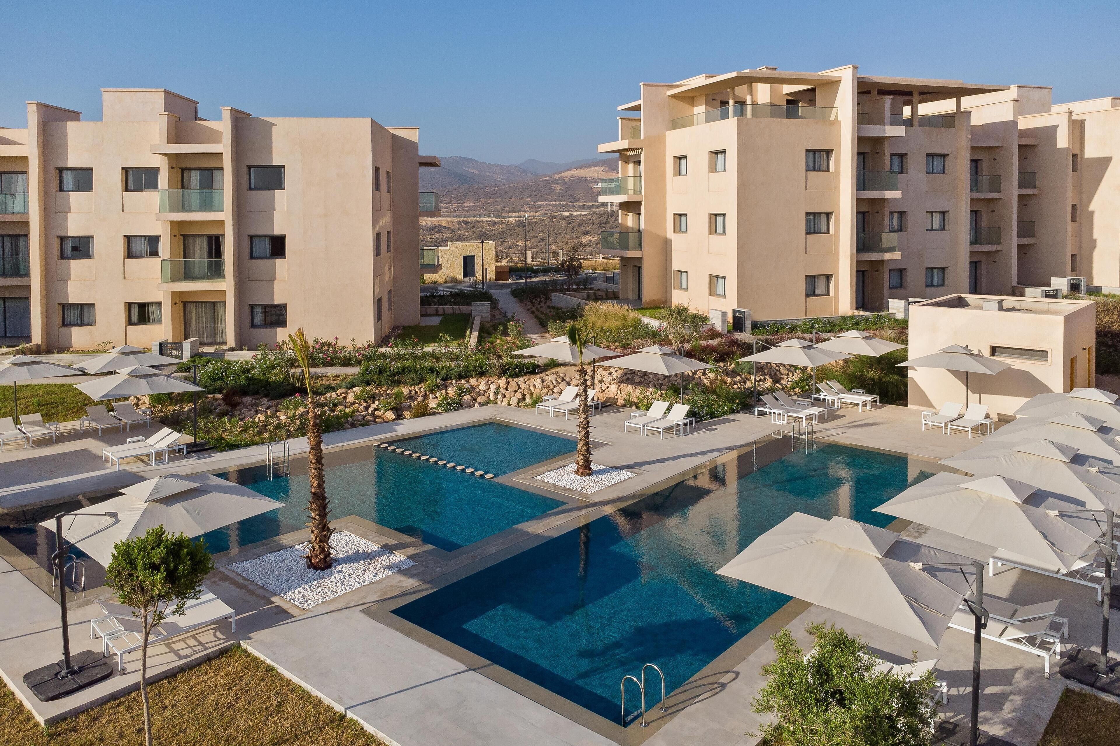 A modern hotel with a central swimming pool, multiple balconies, and outdoor seating areas.