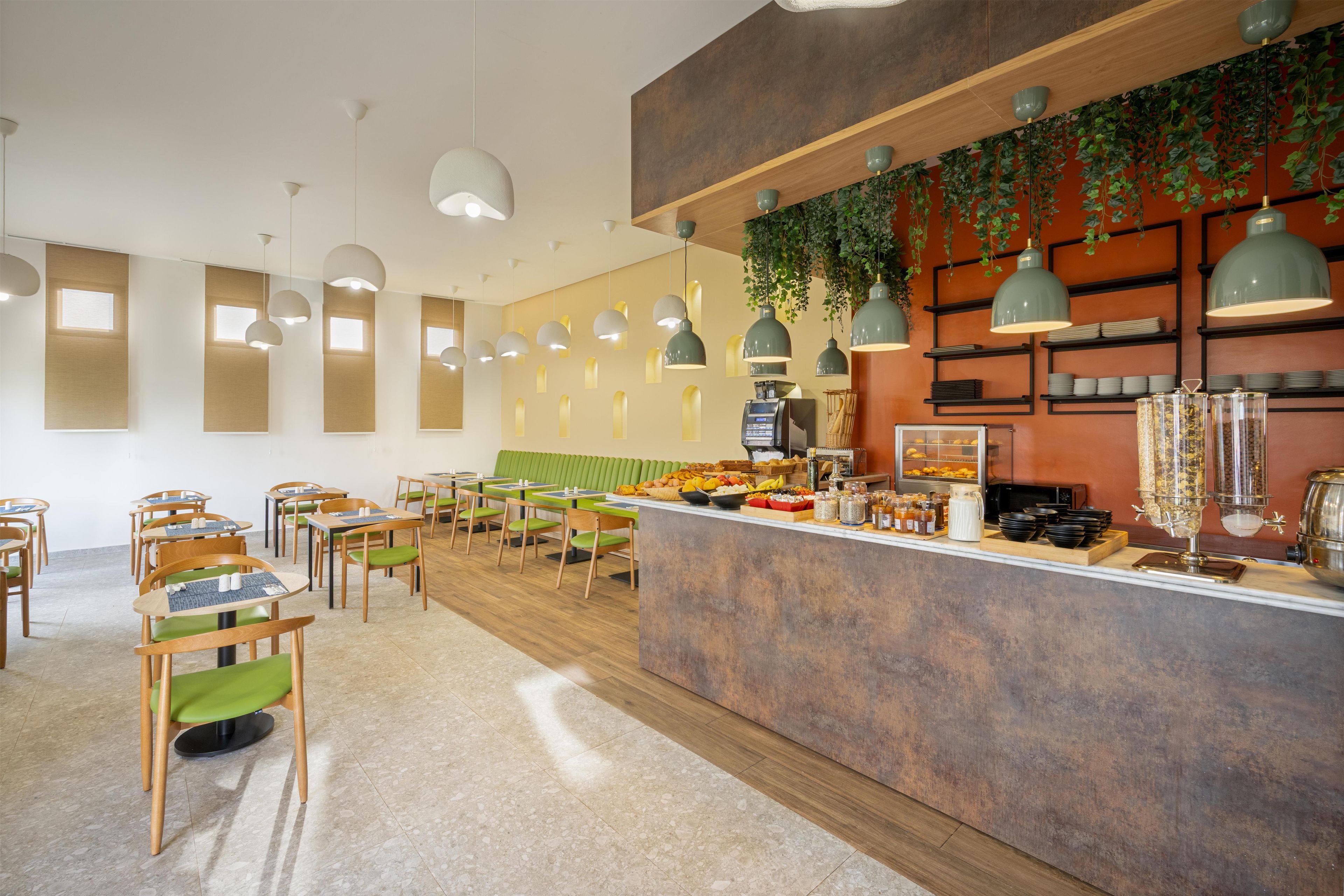 Un café con mesas y sillas de madera, un mostrador con opciones de comida y bebida, y plantas colgantes.