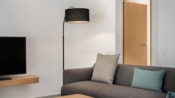 Una sala de estar moderna con un televisor de pantalla plana sobre una estantería de madera, una lámpara de pie oscura, un sofá gris con dos cojines y una mesa de centro de madera.