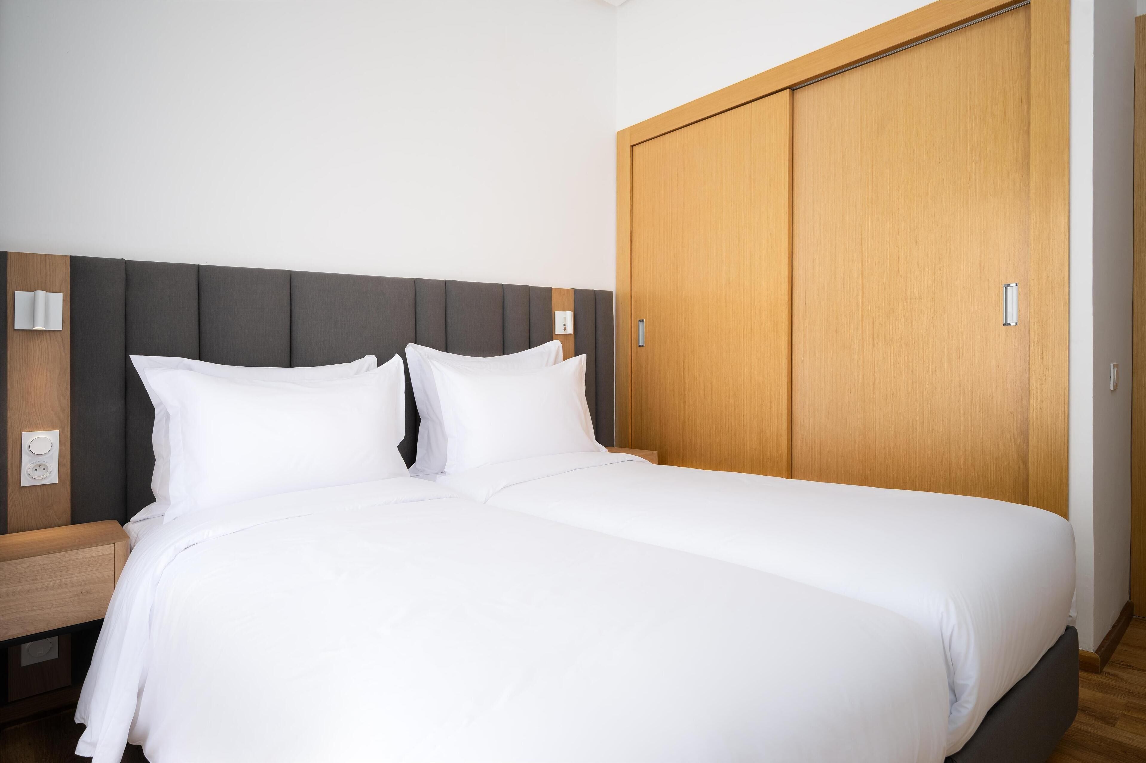 A neatly made bed with white linens, a wooden wardrobe, and a bedside table with a lamp.