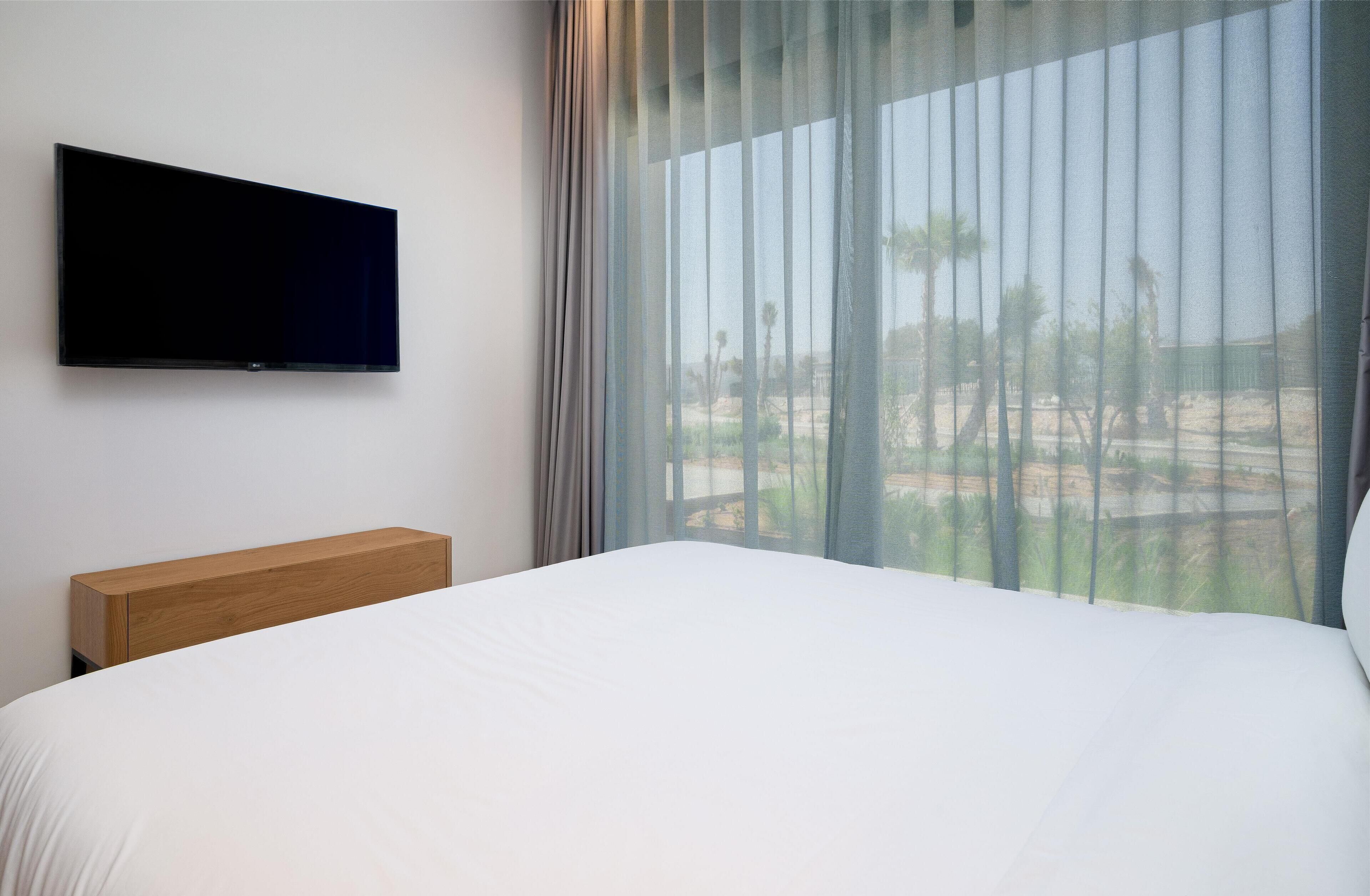 A hotel room with a bed, a wooden headboard, a flat-screen TV mounted on the wall, and a large window with sheer curtains.