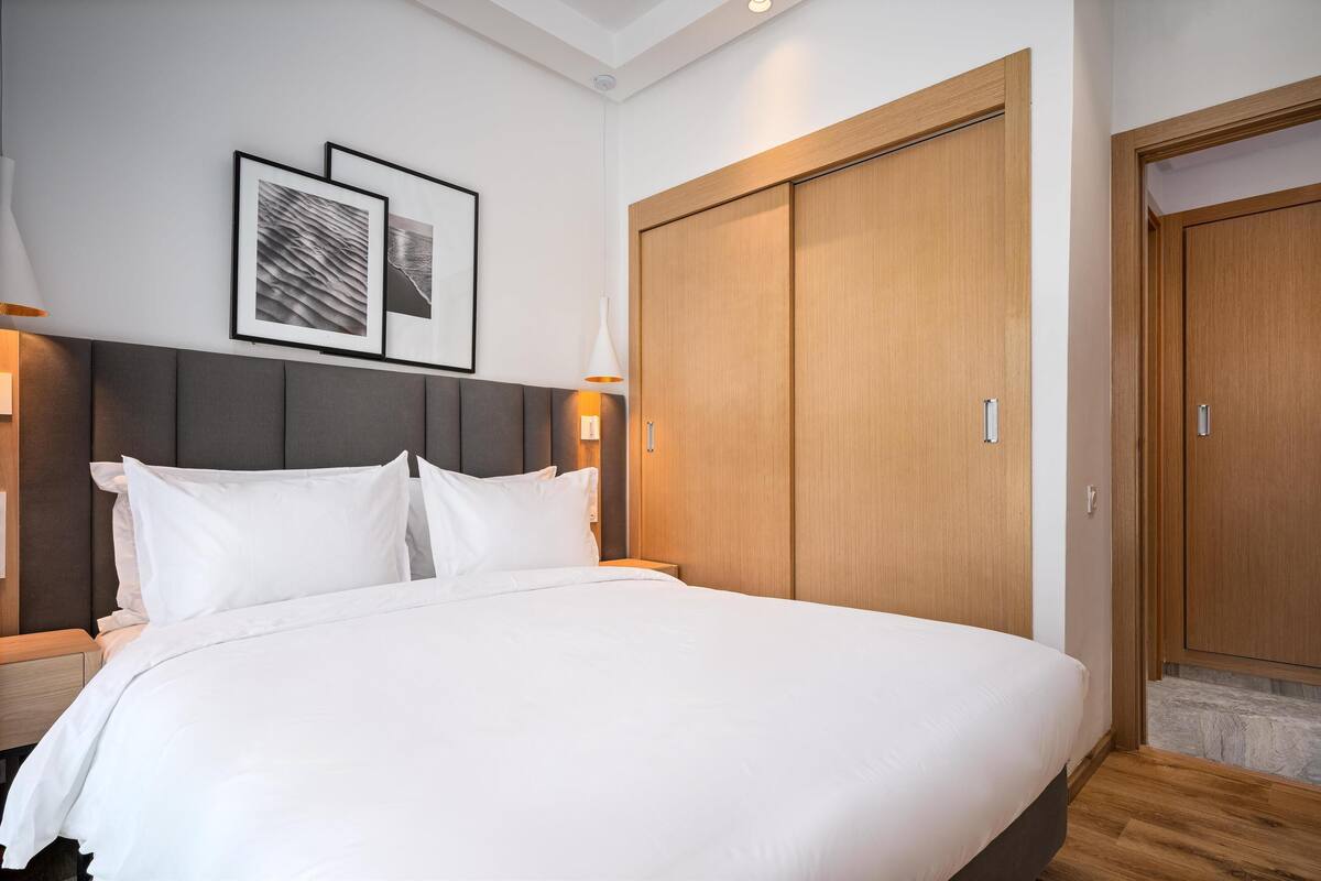 A modern hotel room with a large bed, a grey headboard, wooden wardrobe, and framed artwork on the wall.