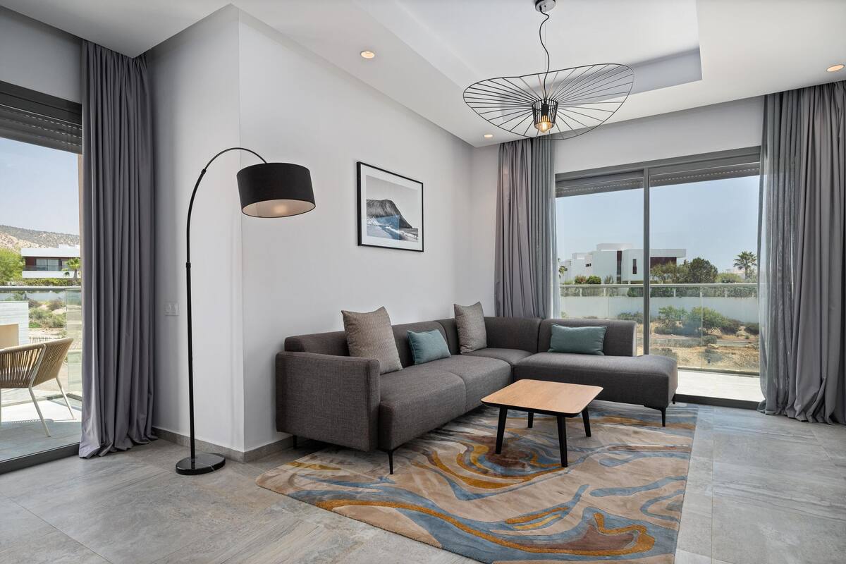 A modern living room with a sectional sofa, a floor lamp, and a large window offering a view of the city.