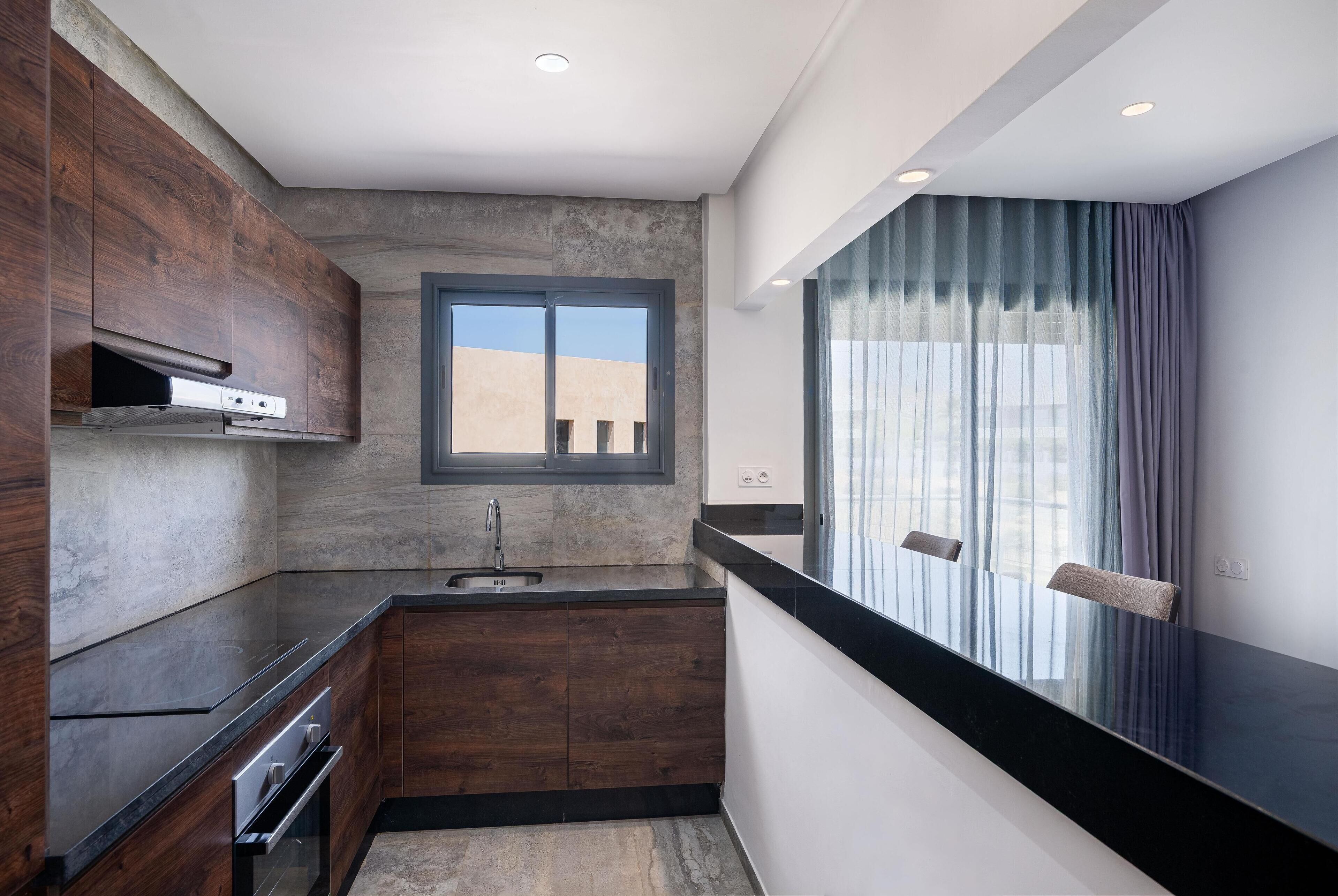 A modern kitchen with dark wood cabinets, a black countertop, and a window with sheer curtains.