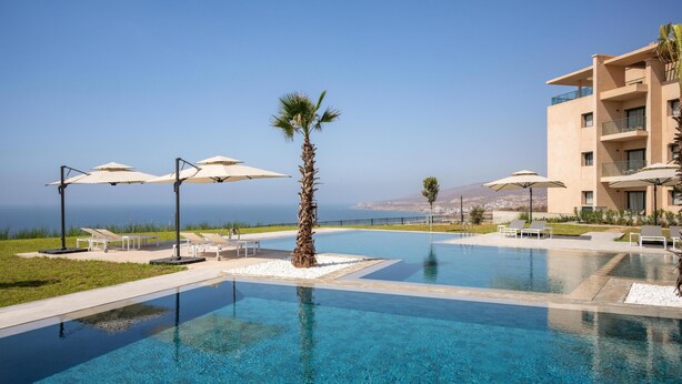 A clear blue pool with lounge chairs and umbrellas, surrounded by a building with balconies and a view of the sea. - Radisson Residences Taghazout Bay (Taghazout)