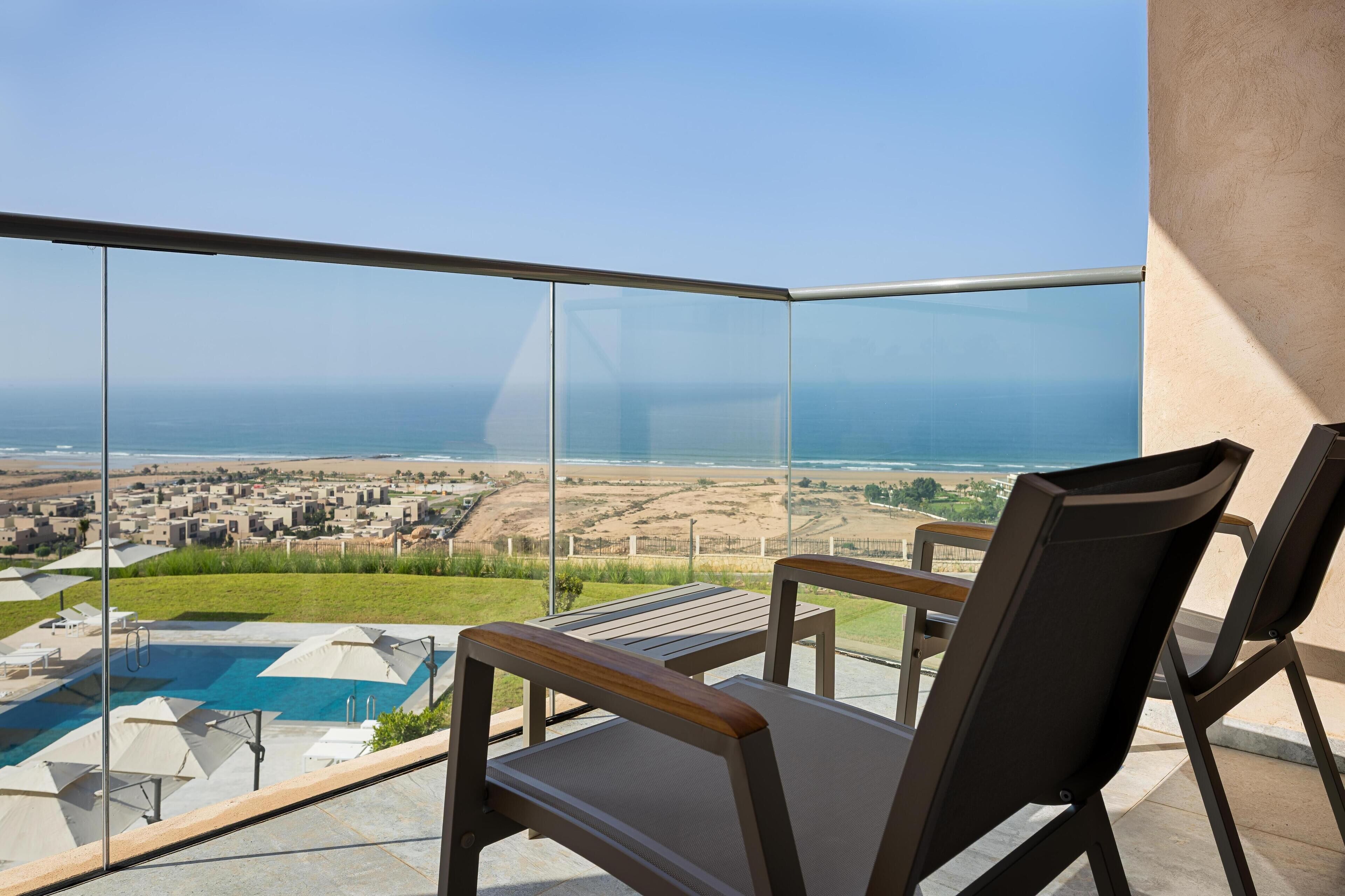 Balcón con barandilla de vidrio que da a una piscina y vistas a la playa.