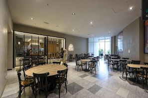 Een ruime eethoek met meerdere tafels en stoelen, een keuken zichtbaar door een glazen scheidingswand, en grote ramen die veel daglicht binnenlaten.