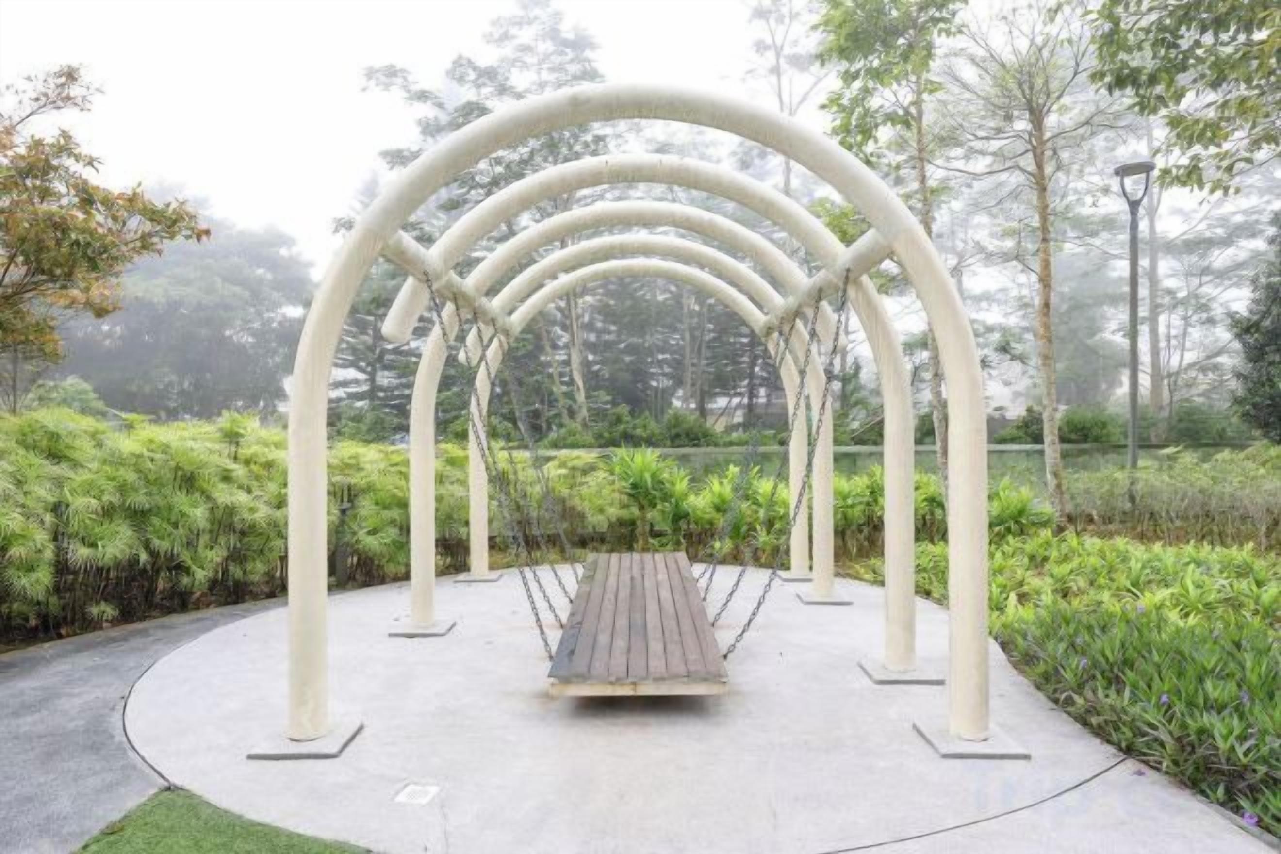 A swing set with a wooden seat and white metal frame, surrounded by greenery and trees.