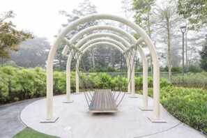 Un columpio con asiento de madera y estructura metálica blanca, rodeado de follaje y árboles.