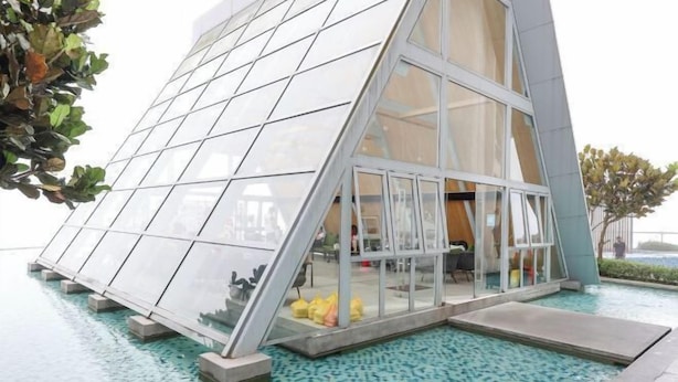 A modern building with a triangular glass facade, a pool area with blue mosaic tiles, and a view of the water.