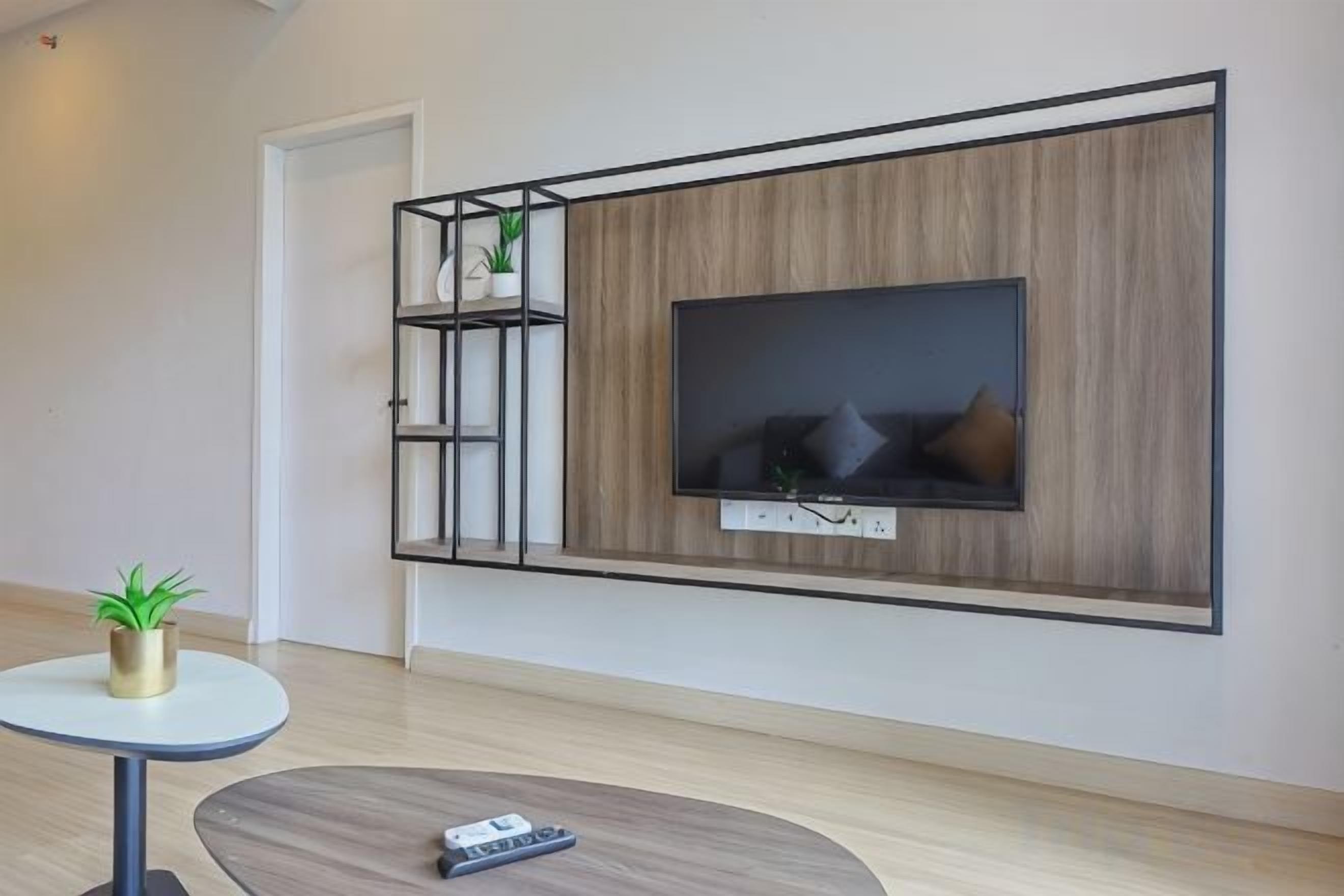 Un salon moderne avec un téléviseur fixé au mur, une petite table ronde sur laquelle se trouve une télécommande, et une plante posée sur une table blanche.