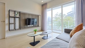 A modern living room with a flat-screen TV, a small table with a plant, a sofa, and a large window with a view of buildings.
