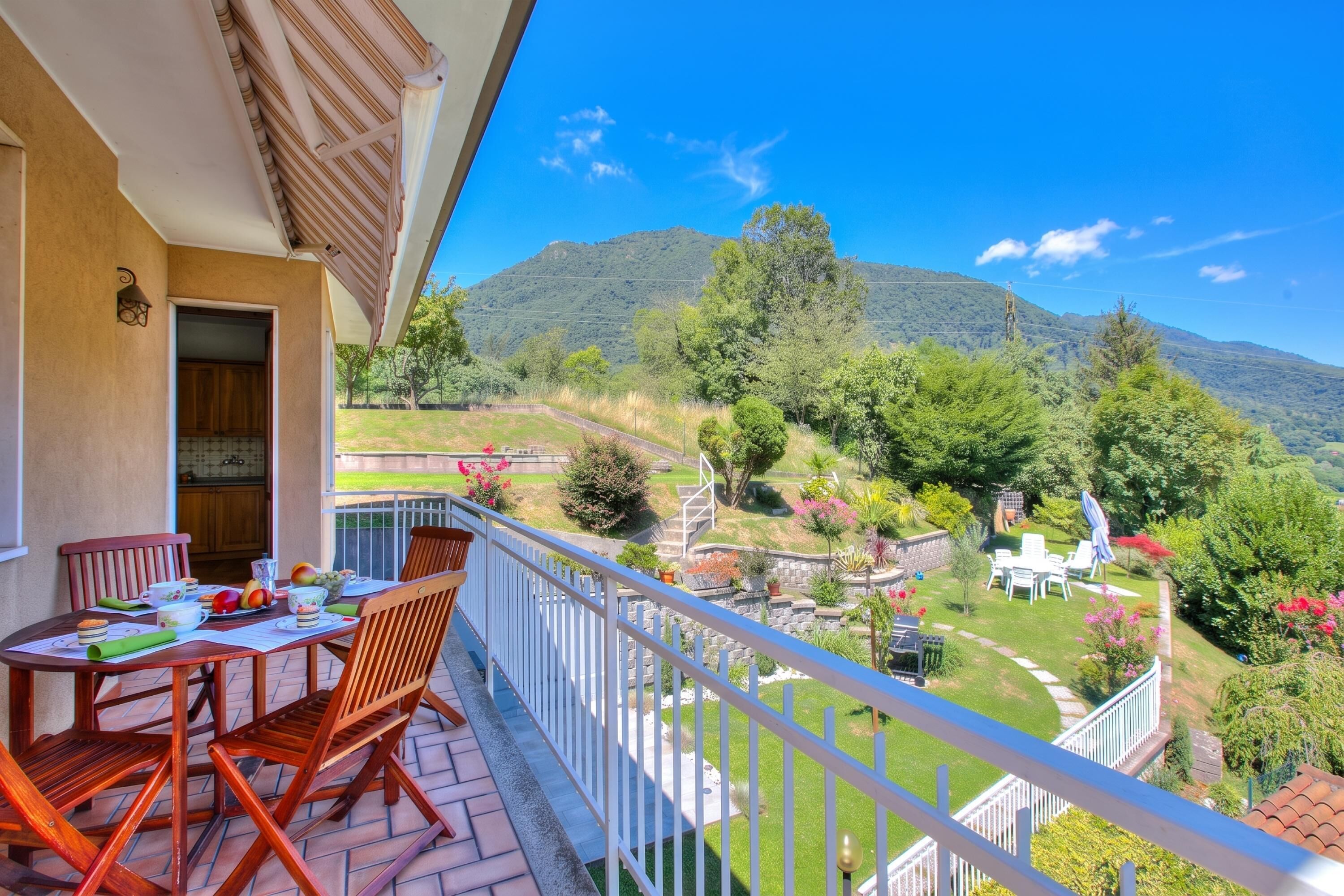 Un balcon avec une table dressée pour deux personnes, donnant sur un jardin aménagé et un paysage montagneux.